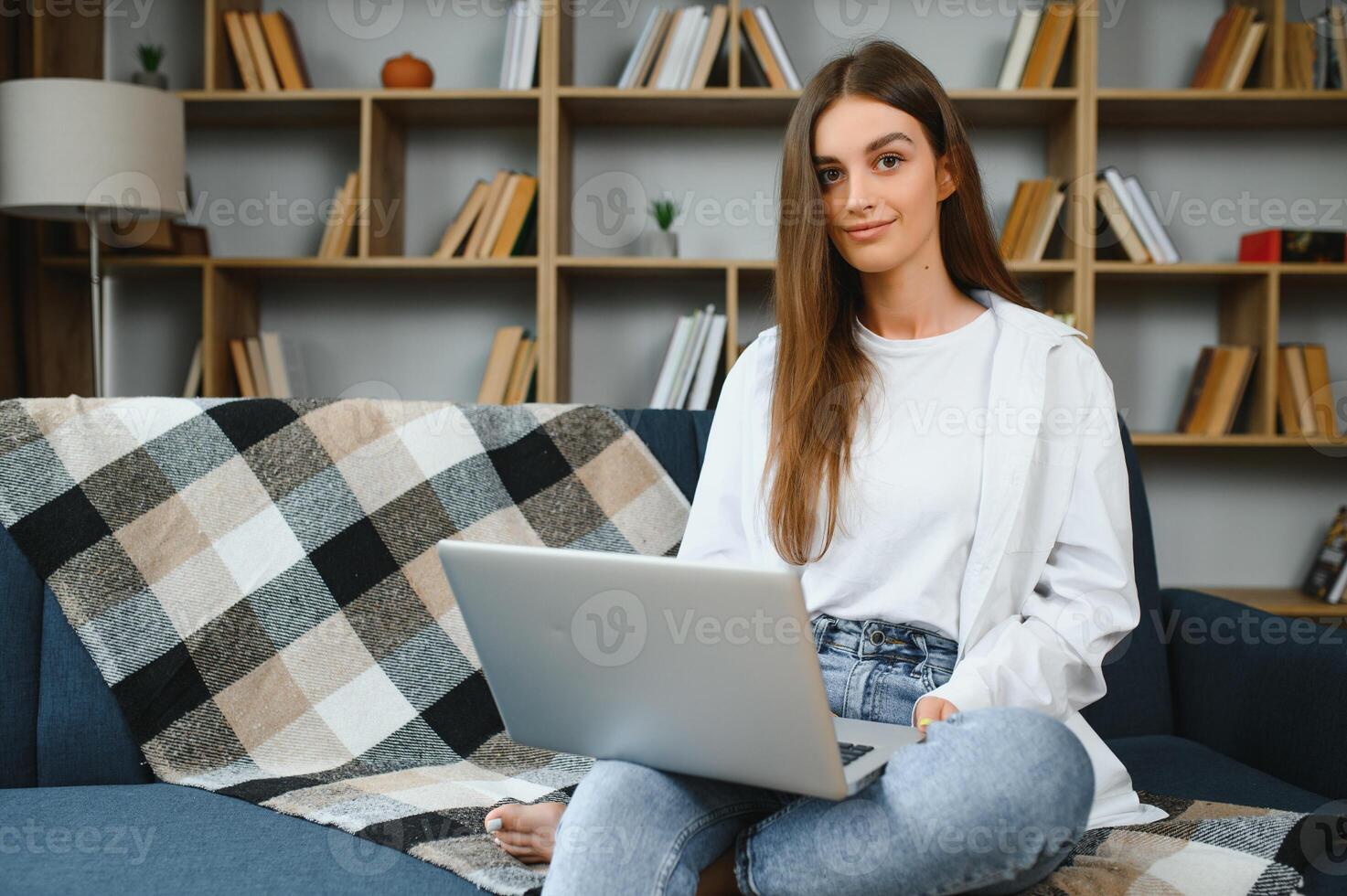 Smiling attractive young woman sitting on sofa using laptop communicating working online at home, happy teen girl typing on computer, enjoying writing blog or chatting with friends in social network photo