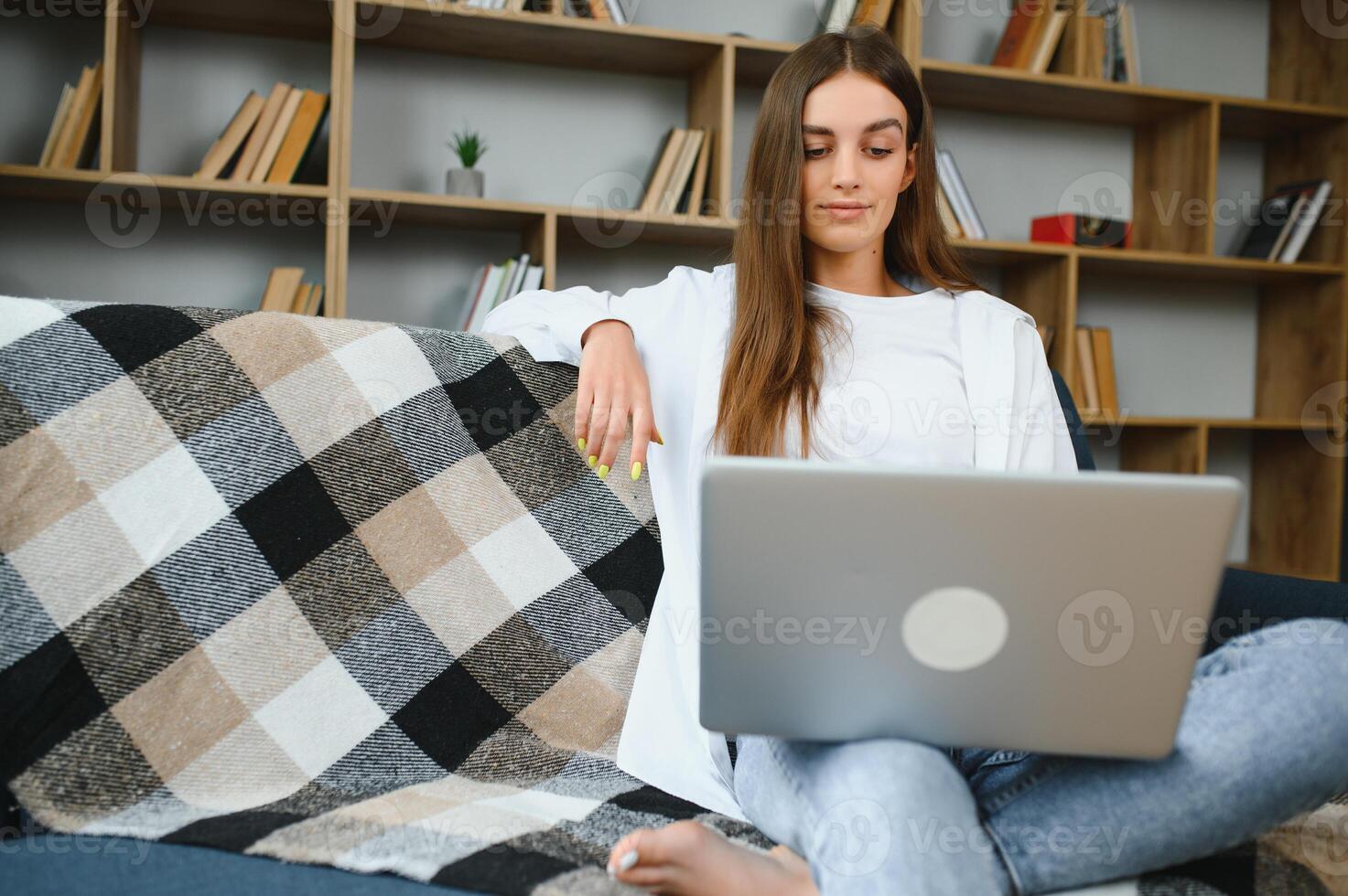 Smiling attractive young woman sitting on sofa using laptop communicating working online at home, happy teen girl typing on computer, enjoying writing blog or chatting with friends in social network photo