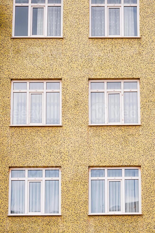 Facade of a yellow textured building with six white framed windows arranged in two columns and three rows. photo