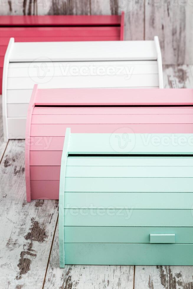 Colorful wooden bread boxes placed on a rustic white wooden surface. Bread boxes featuring a roll top opening mechanism with horizontal slats. photo