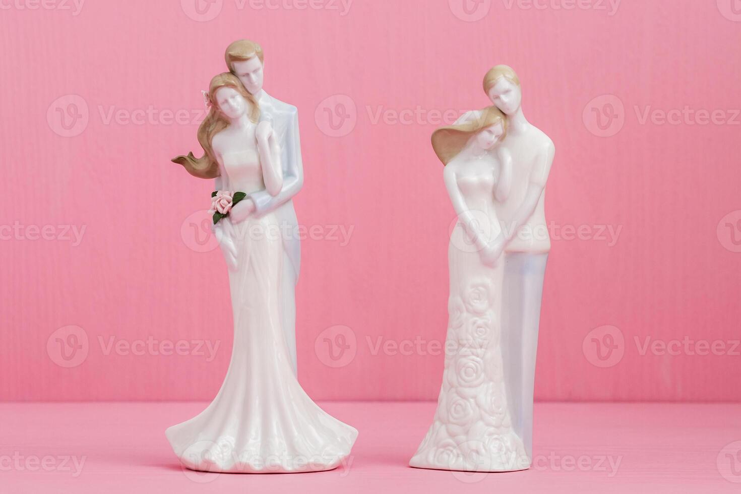 Two porcelain wedding figurines of couples embracing, set against a pink background with one couple holding a bouquet of flowers. photo