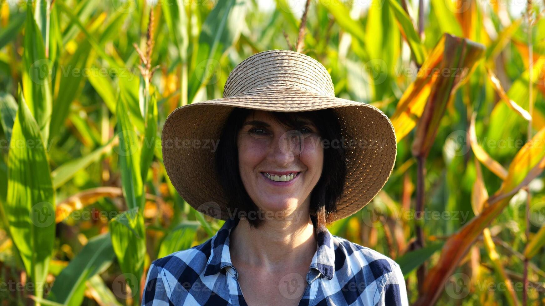 Happy smiling female farmer looks into camera standing near corn field. Portrait of adult beautiful agronomist in straw hat with maize meadow at background. Agricultural business concept. Close up photo