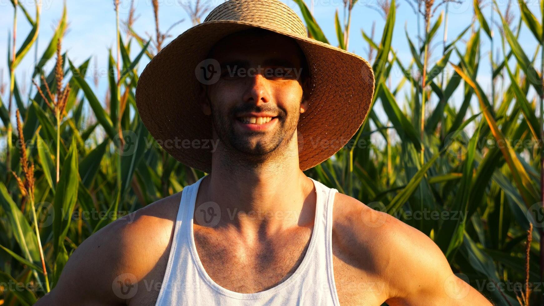 Happy smiling male farmer looks into camera standing near corn meadow. Portrait of young handsome agronomist in straw hat with maize field at background. Agricultural business concept photo