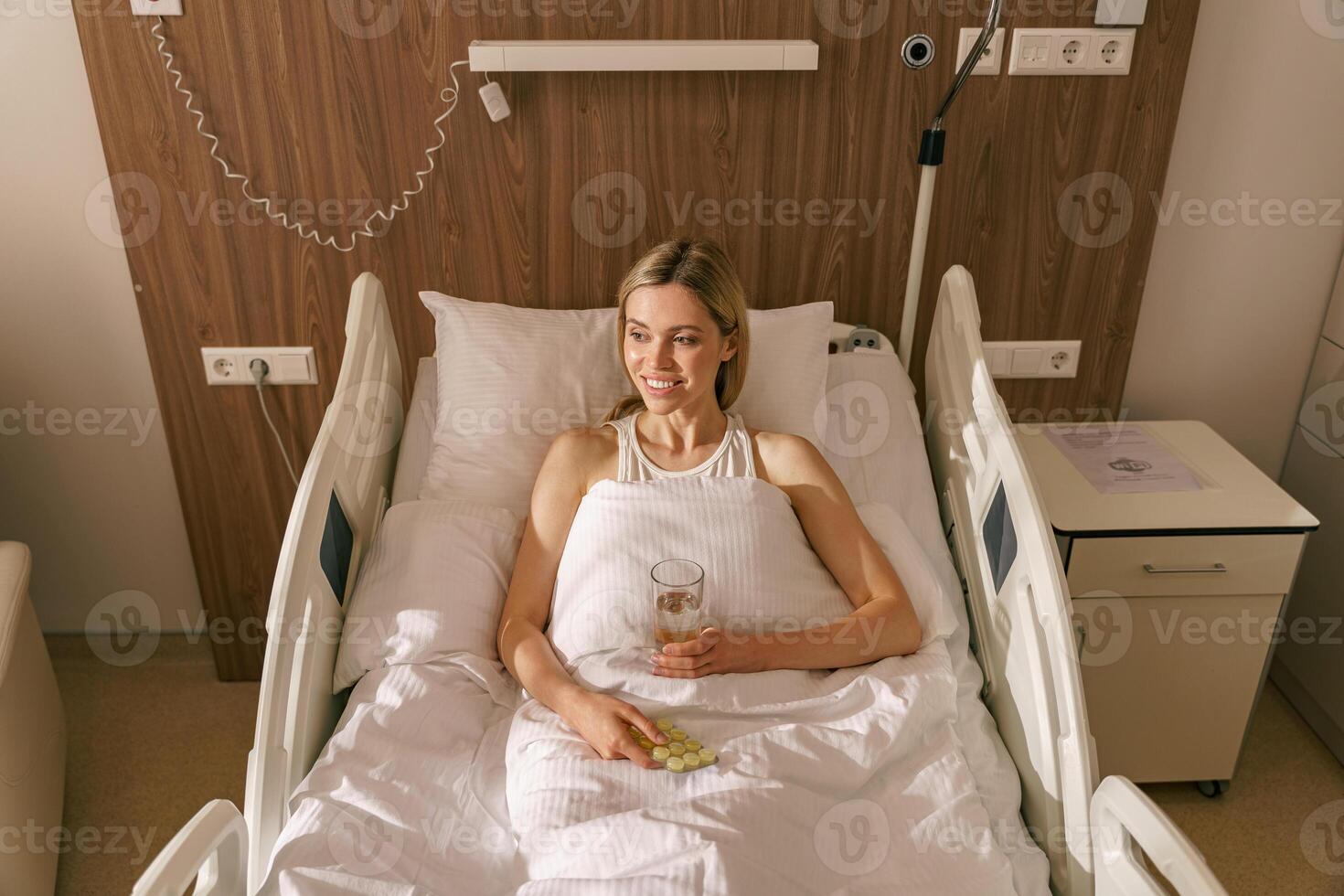 Woman patient lying in hospital ward and holding pills and glass of water while looking at side photo