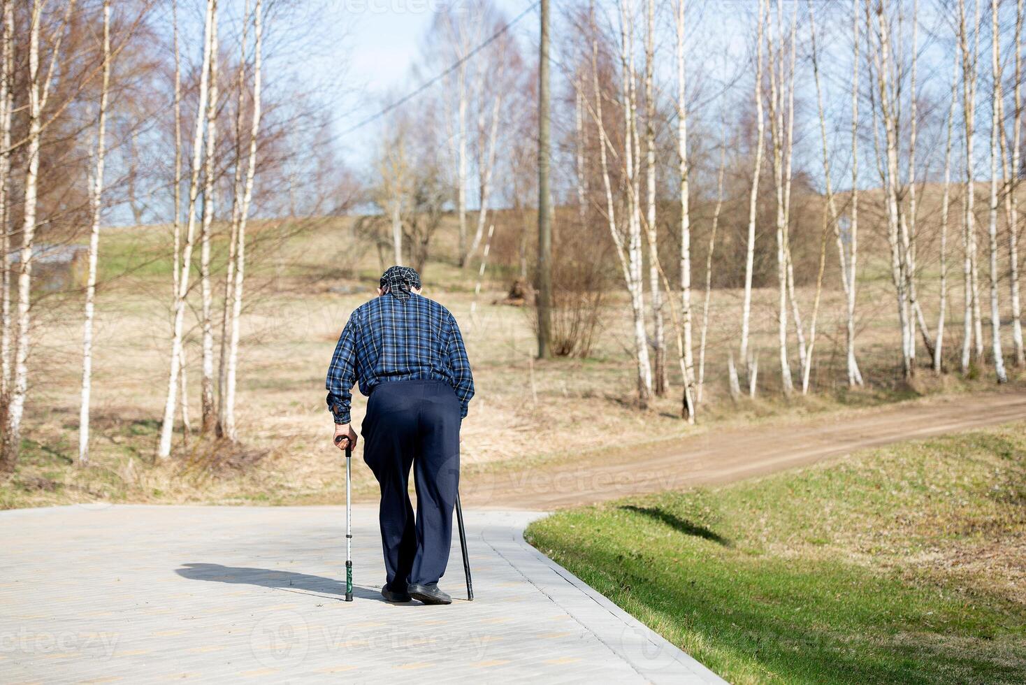 An old man wearing a headscarf and sunglasses walks outdoors leaning on two sticks photo