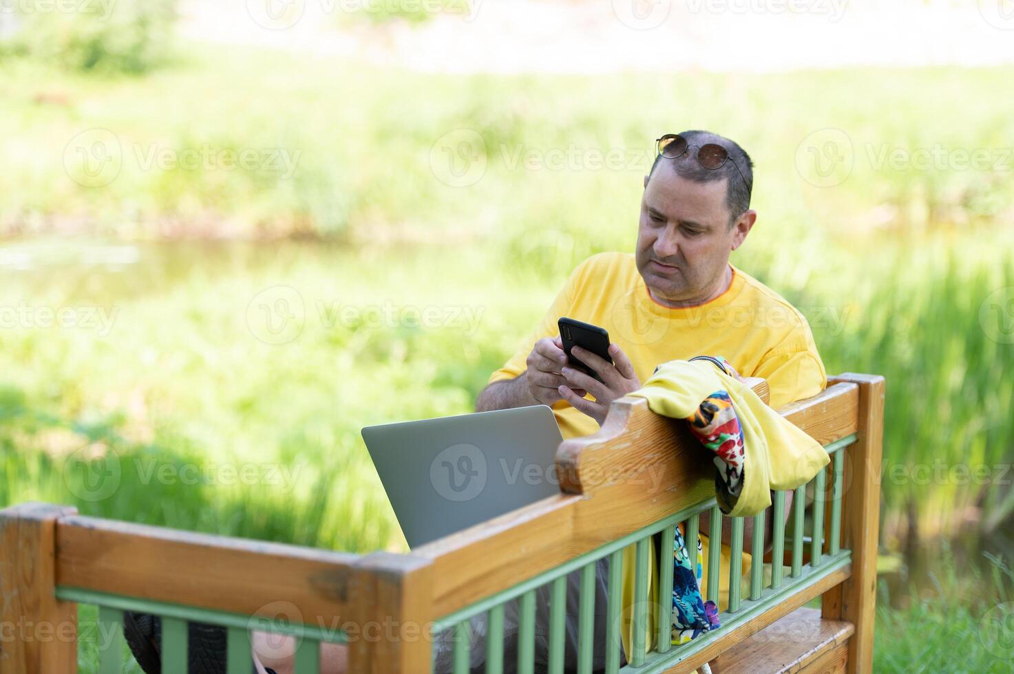 european man 50s in casual clothing sitting on bench in park and talking on cell phone while using silver laptop photo