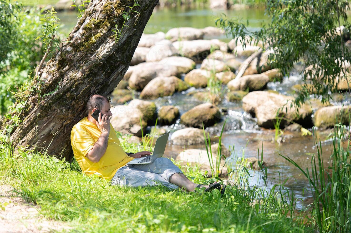 Middle aged freelancer using a laptop computer and phone while working in beautiful nature under the tree on the bank of the river photo