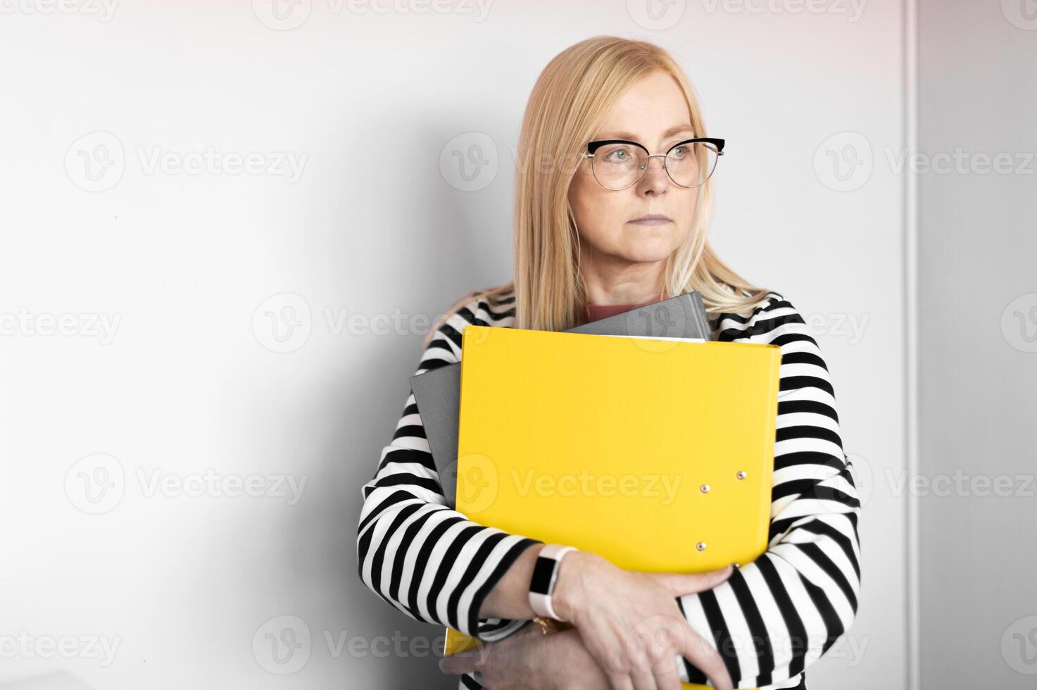 middle aged blonde business woman with glasses and yellow document folder isolated on light office wall background photo