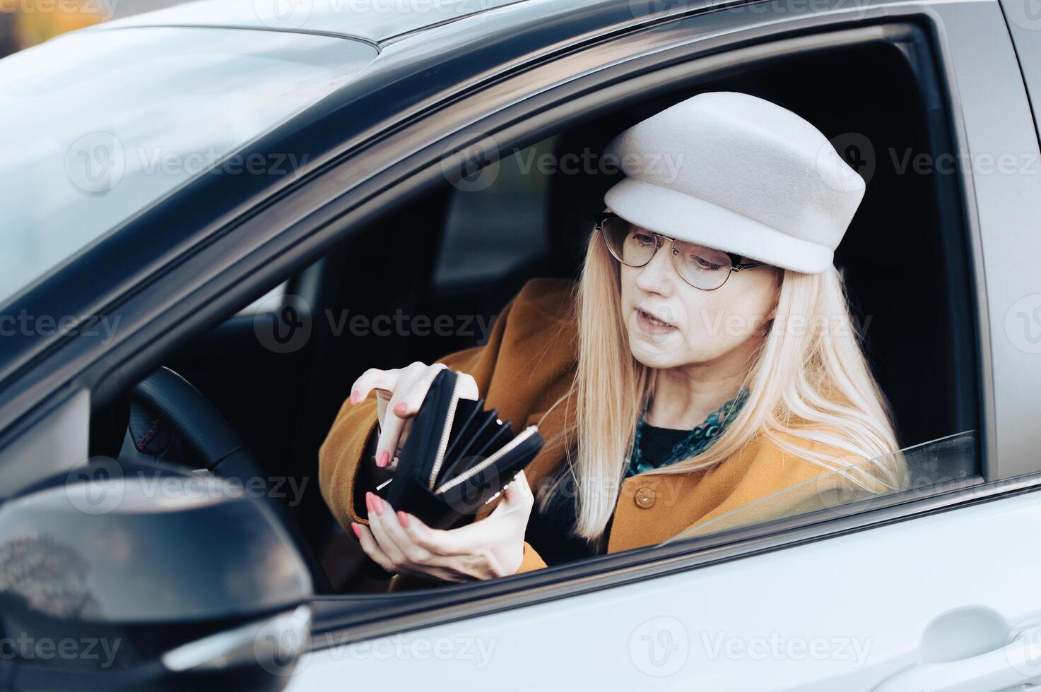 Middle-aged woman sits behind the wheel of car and looks for money in wallet to pay for parking photo