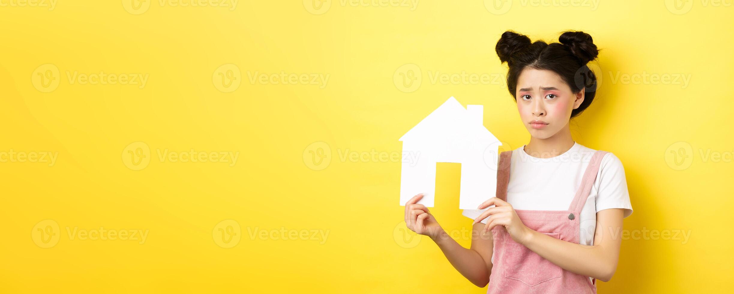 Real estate and family concept. Sad cute asian girl with bright makeup, frowning and feel upset, showing paper house cutout, standing on yellow background photo