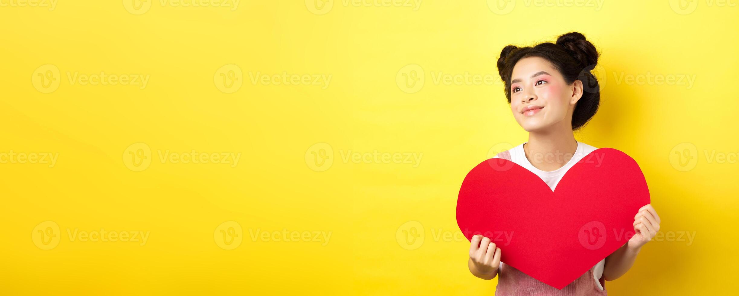 Asian girl in love, looking dreamy left and smiling, showing Valentines red heart cutout, imaging romantic date, standing on yellow background photo