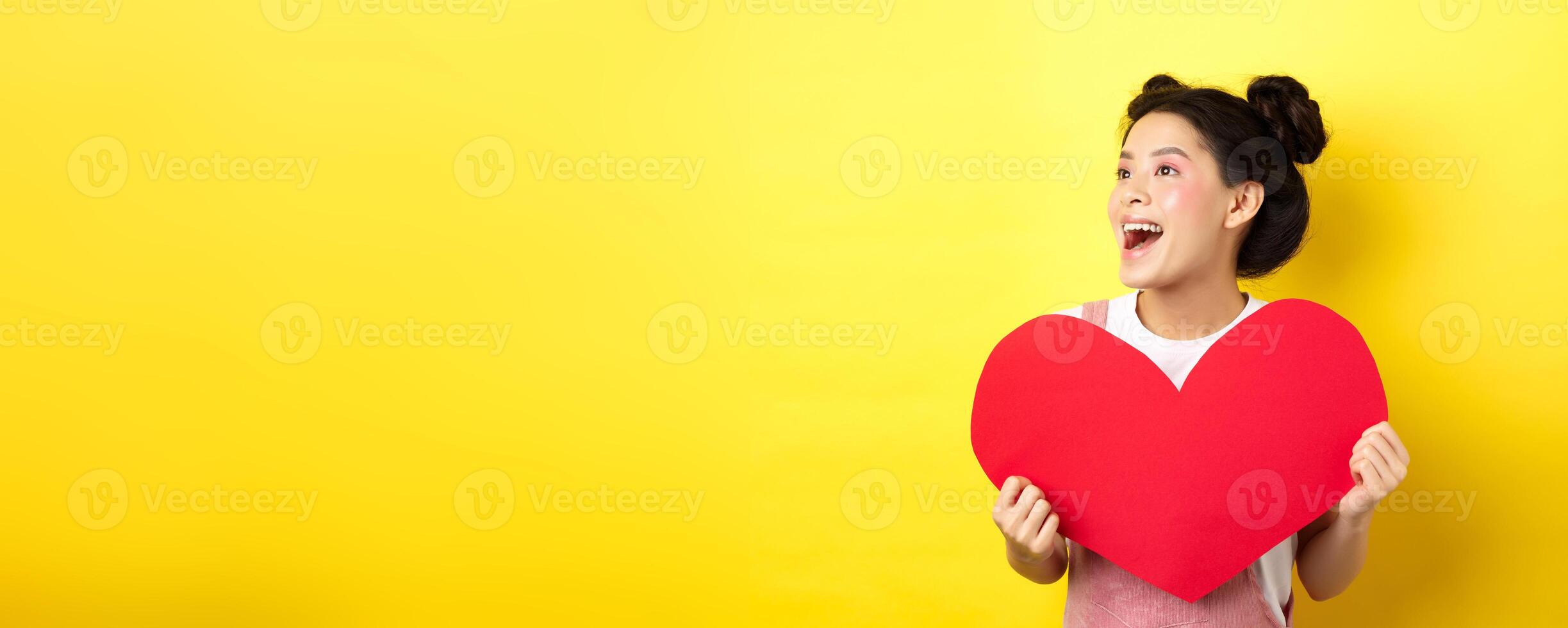 Happy valentines and love concept. Cheerful japanese girl looking left, showing big red heart and smiling happy at lover, standing on yellow background photo