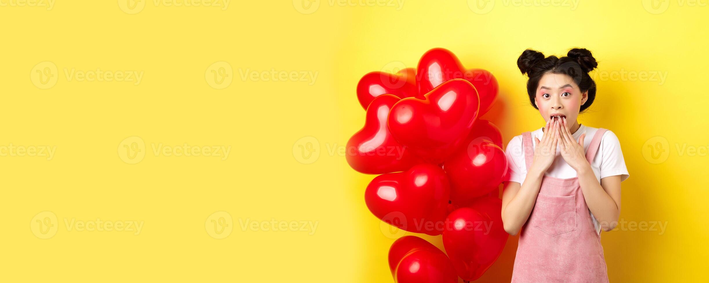 Surprised korean girl gasping and saying wow, looking at special promo offer on Valentines day, standing near big red hearts balloons and yellow background photo