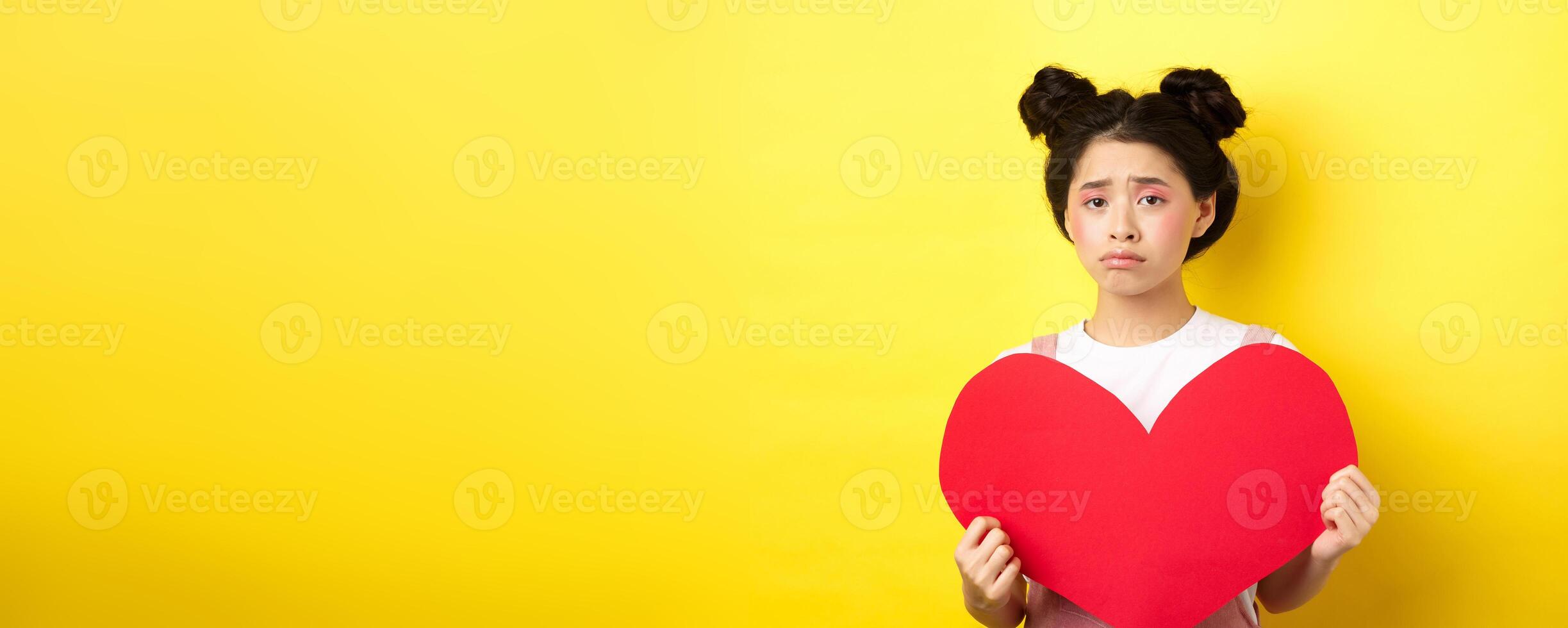 Heartbroken asian woman showing big red heart cutout and looking sad, feeling lonely on lovers day, showing valentine card at camera, yellow background photo