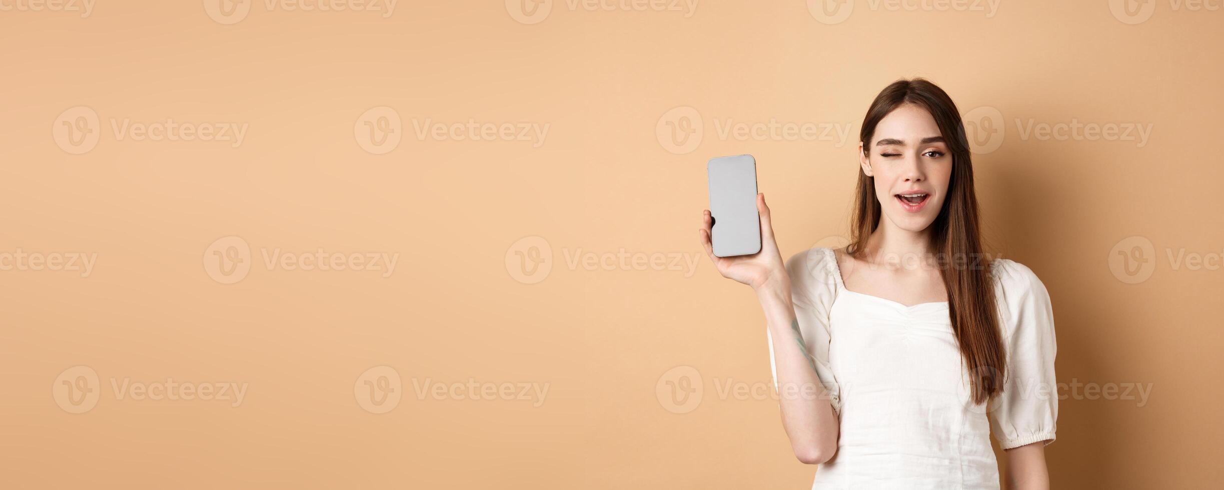 Cheerful stylish girl showing smartphone app, demonstrate empty phone screen and winking, standing on beige background photo