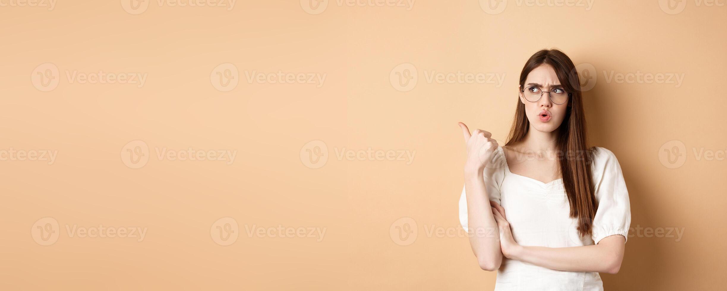 Confused and suspicious young woman in glasses frowning, pointing and looking aside with displeased face expression, standing on beige background photo