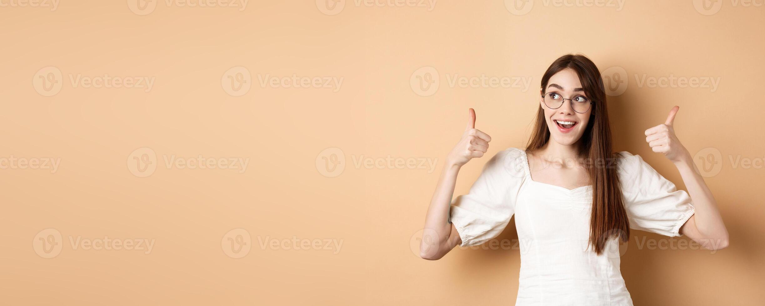 Happy girl in glasses show thumbs up, looking aside at good logo, standing satisfied on beige background photo