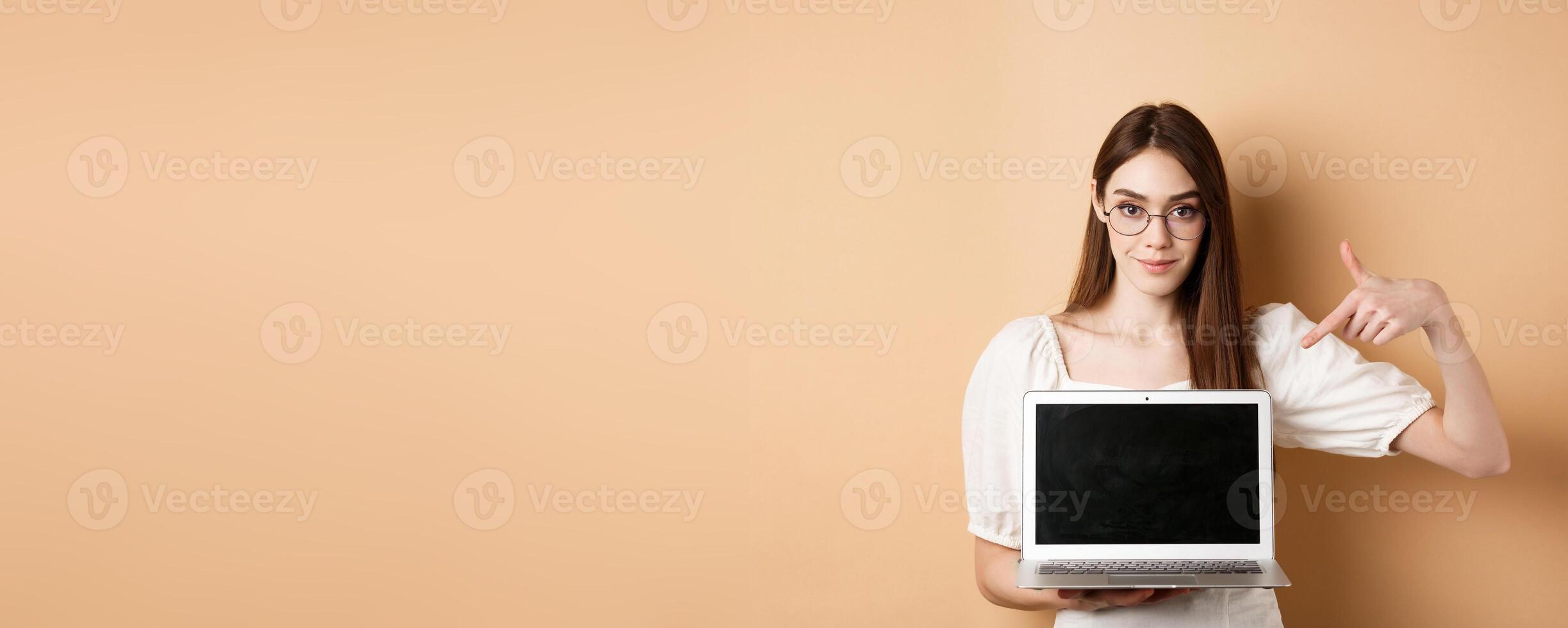 Confident young woman in glasses pointing at laptop screen, showing computer display, standing on beige background photo