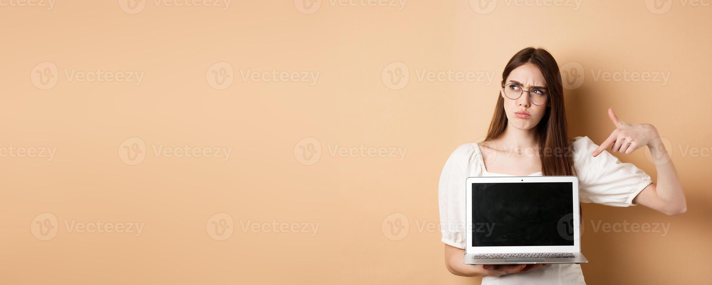 Suspicious frowning girl in glasses pointing at laptop screen, showing something strange online, standing on beige background photo