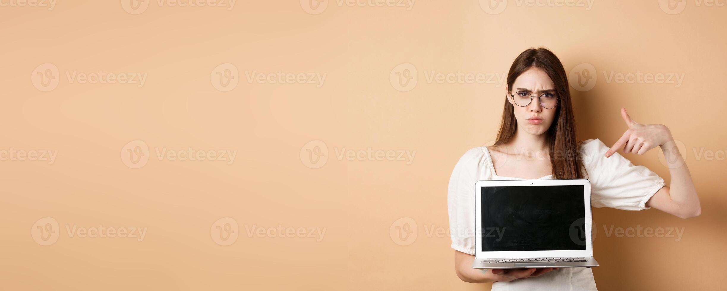 Angry frowning woman in glasses pointing at laptop screen, complaining and look disappointed, standing on beige background photo