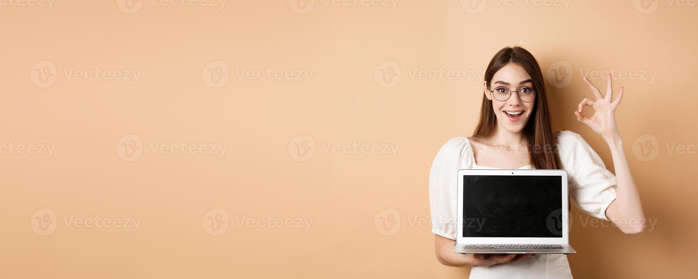 E-commerce. Excited young woman in glasses showing okay sign and laptop screen, recommending internet promo, standing on beige background photo