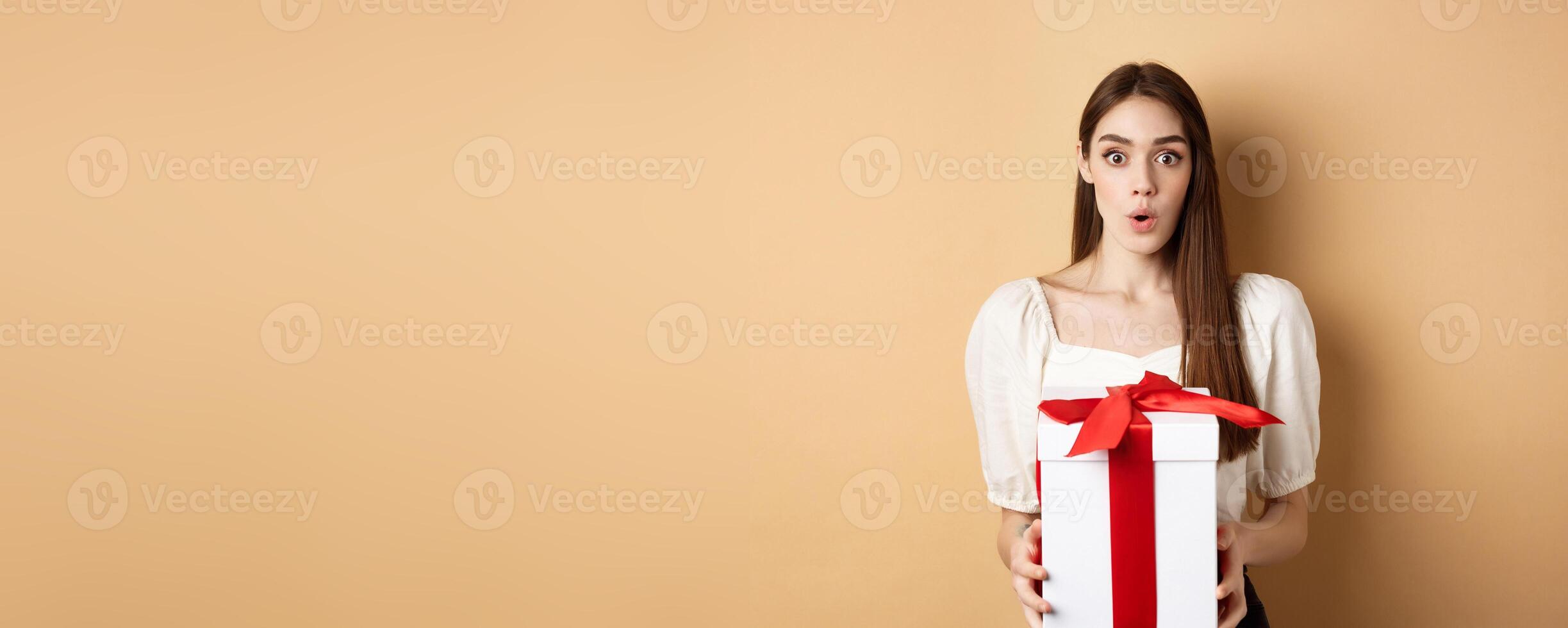 Image of surprised girl receive gift on Valentines day, looking with disbelief and holding present, standing on beige background photo