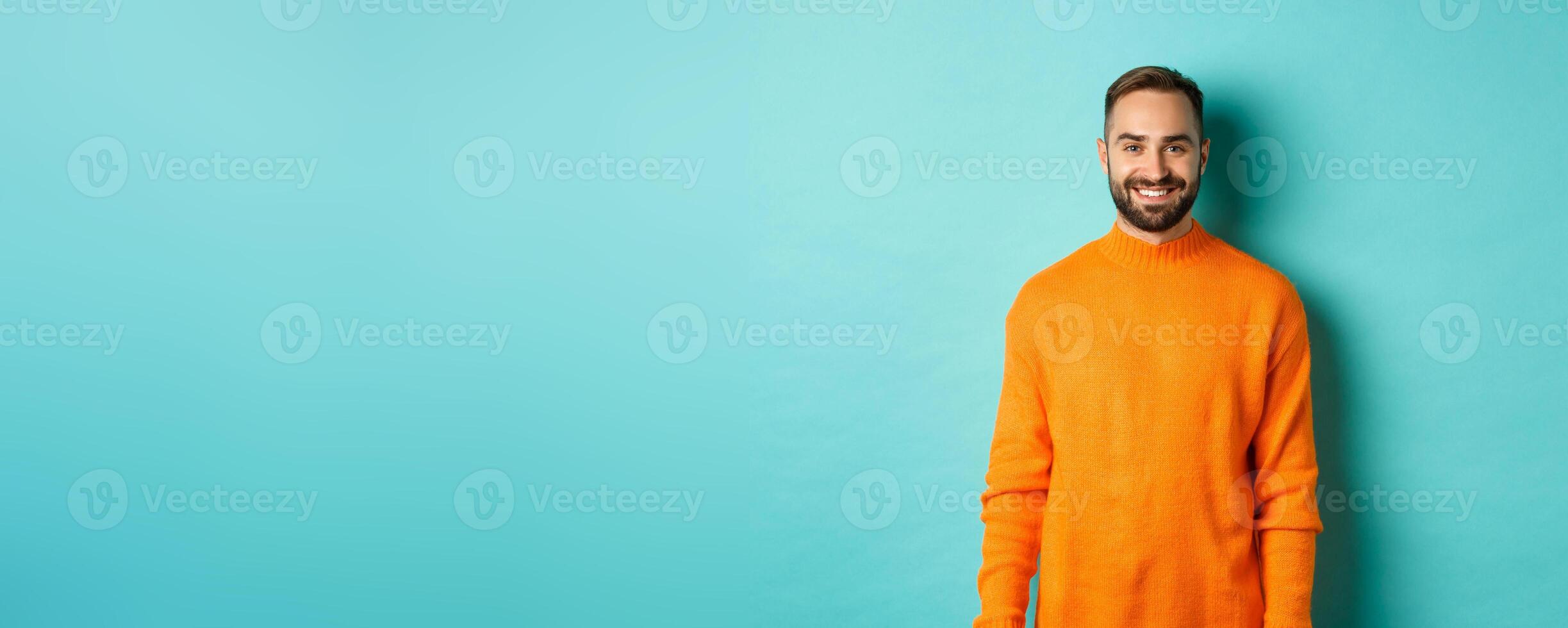 Lifestyle. Handsome man in orange sweater smiling at camera, happy face, guy cheerful expression, standing over turquoise background photo