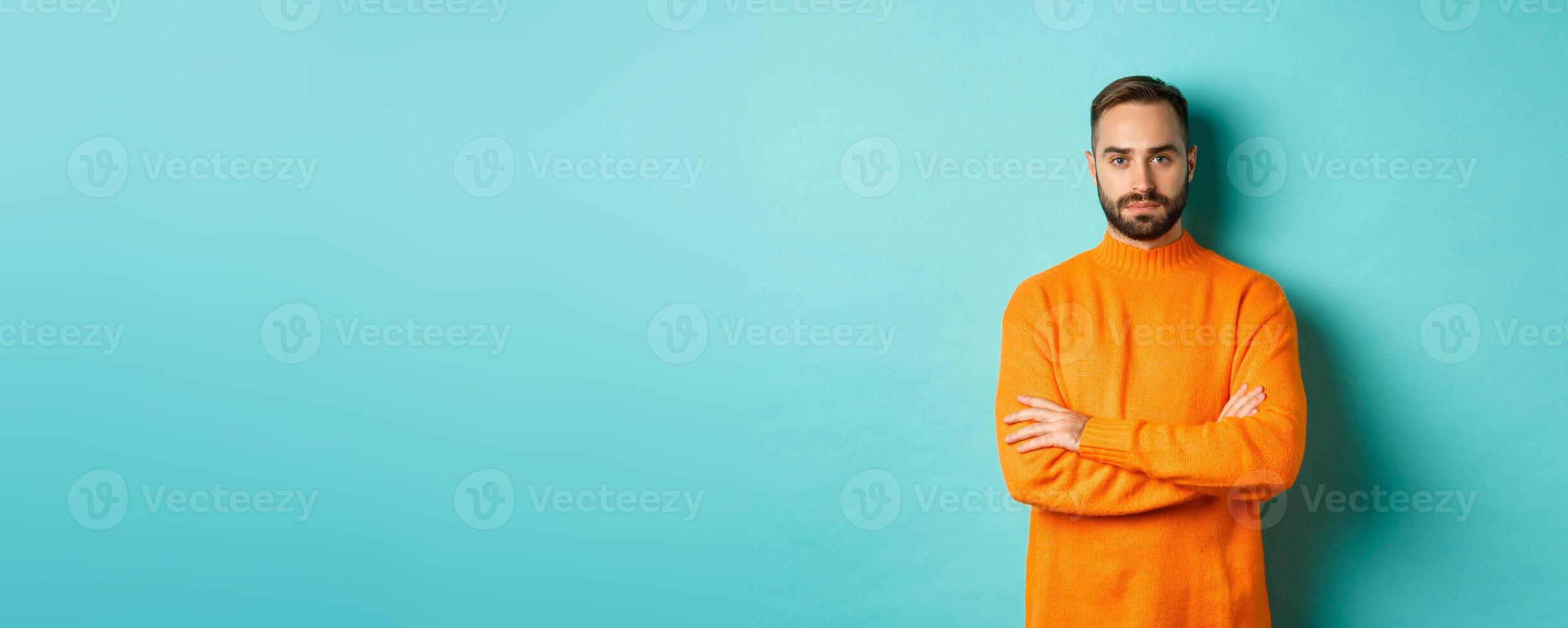 Confident young man looking determined, cross arms on chest, wearing orange winter sweater, standing against turquoise studio background photo