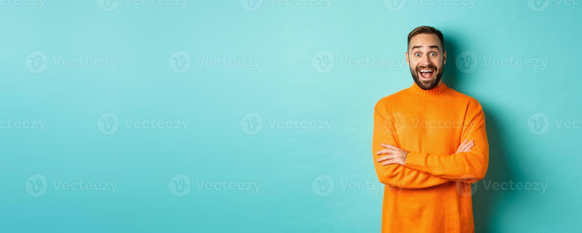 Image of happy and surprised man reacting to news, looking amazed, standing in orange sweater against turquoise background photo