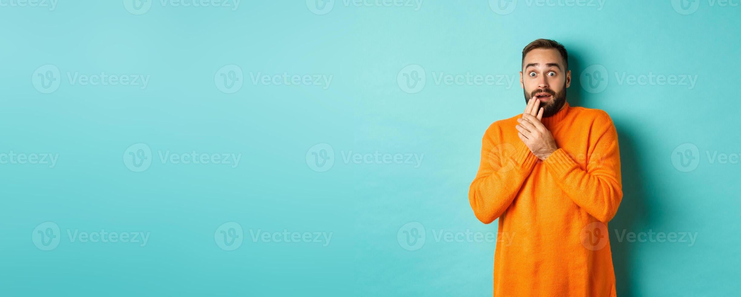 Scared and shocked guy looking at camera, standing in orange sweater against turquoise background photo