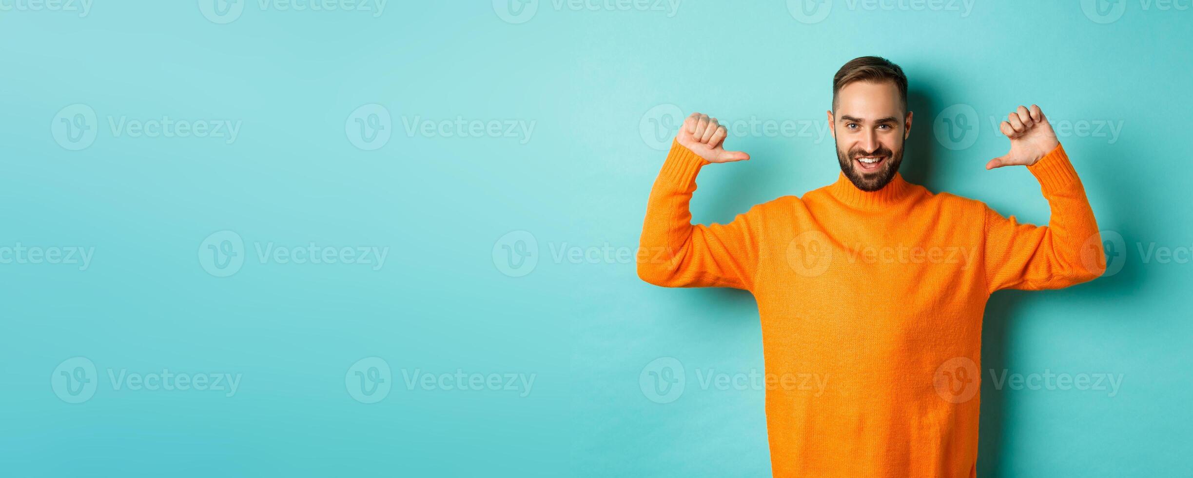 Handsome confident man pointing at himself, looking self-assured, standing in orange sweater against light blue background photo