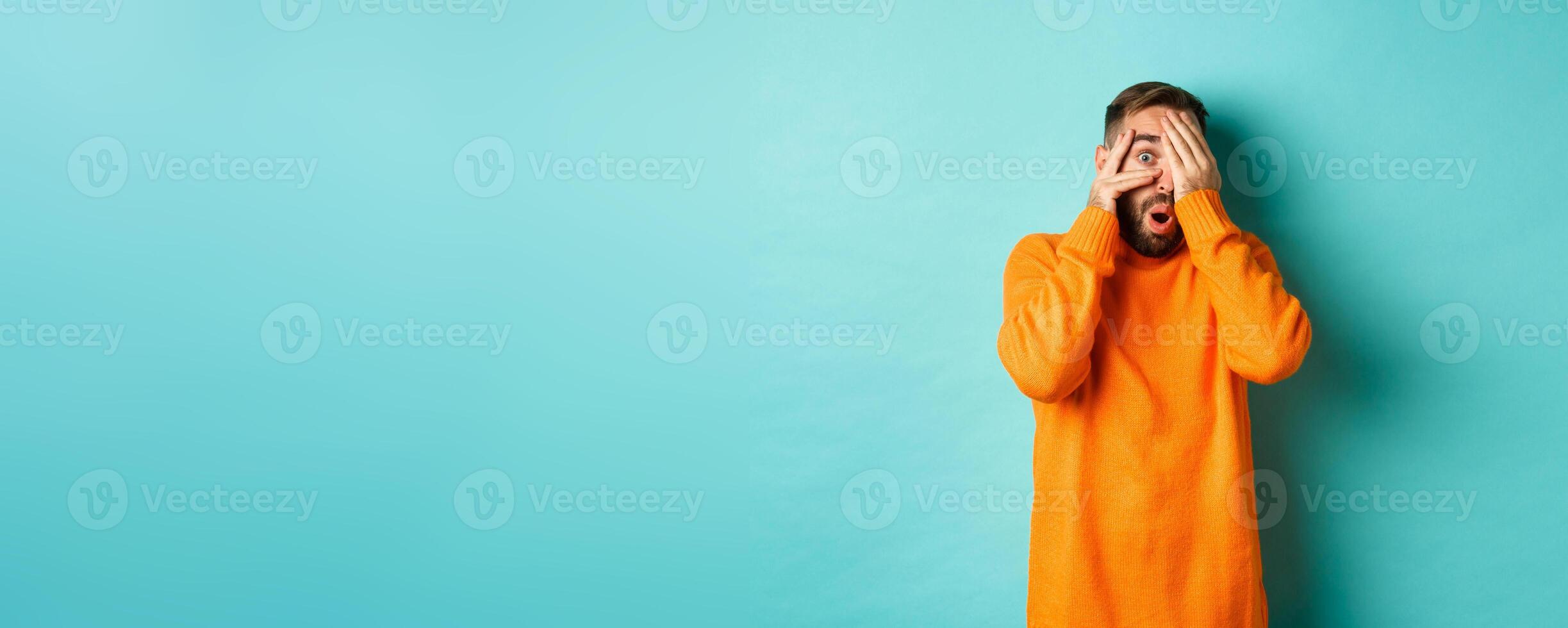 Curious man peeking at something, shut eyes but looking through fingers with amazement and interest, standing over light blue background photo