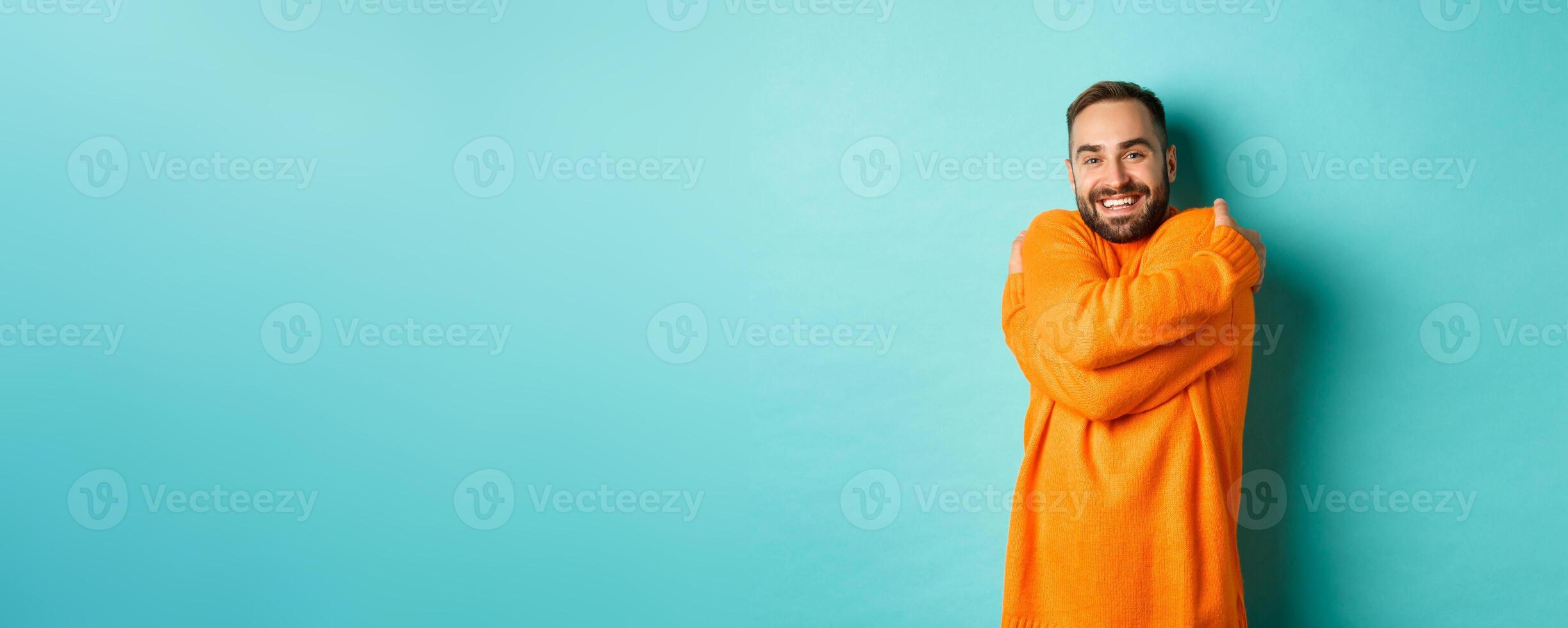 Happy man feeling comfortable, wearing warm sweater and hugging himself, smiling satisfied, standing over light blue background photo