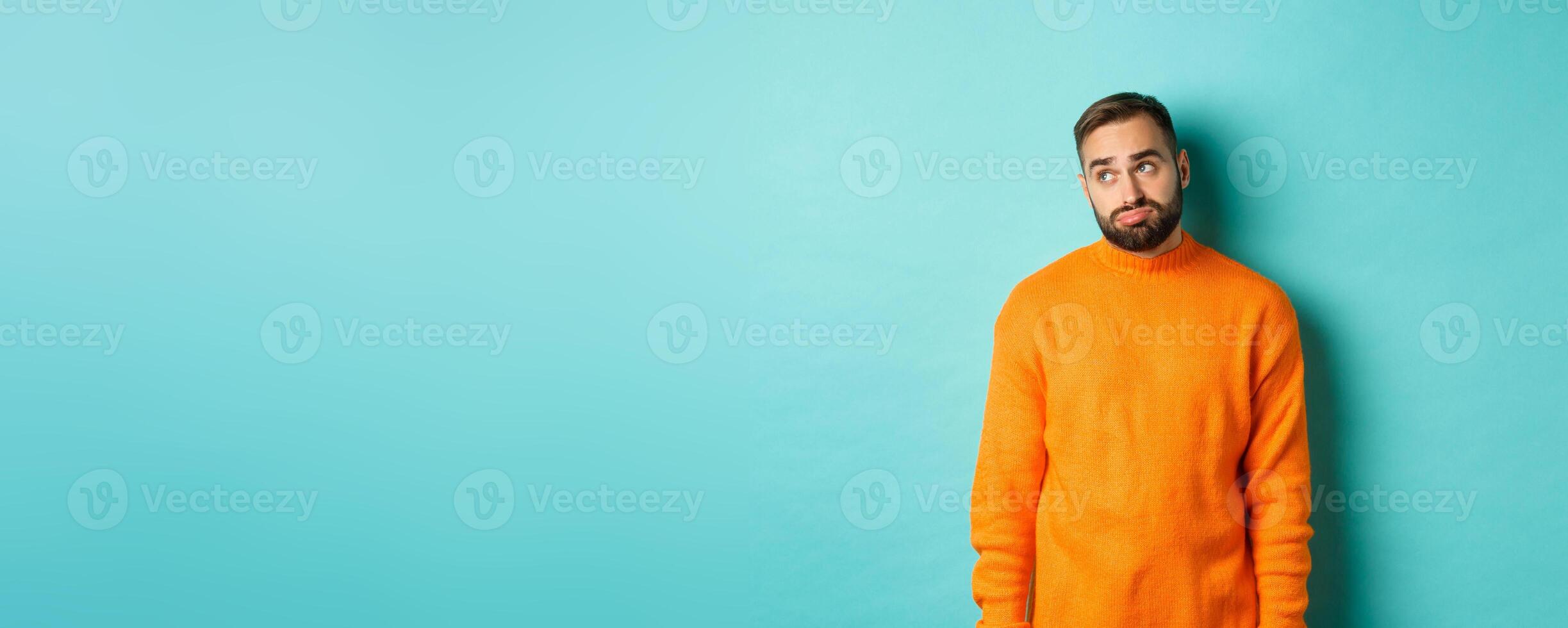 Sad and gloomy man sulking, looking bored at upper left corner, standing in orange sweater against light blue background photo