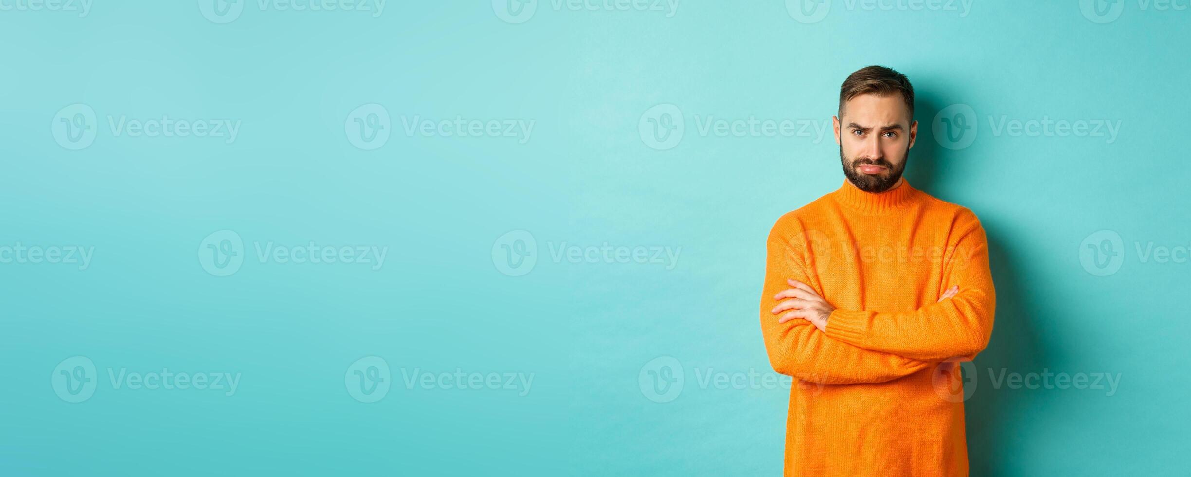 Silly sad guy with beard, sulking and looking offended, cross arms on chest and frowning upset, standing over light blue background photo