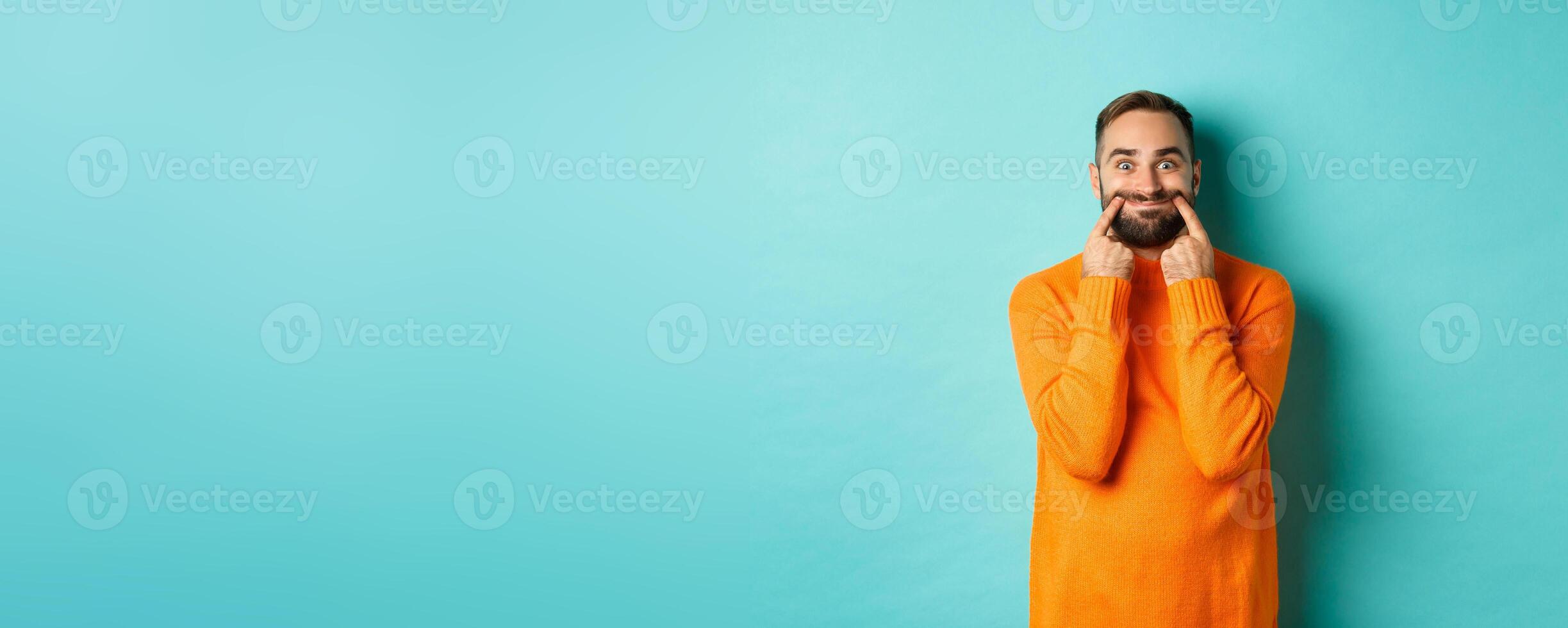 Image of bearded man stretching lips in happy smile, faking happiness, standing over light blue background photo