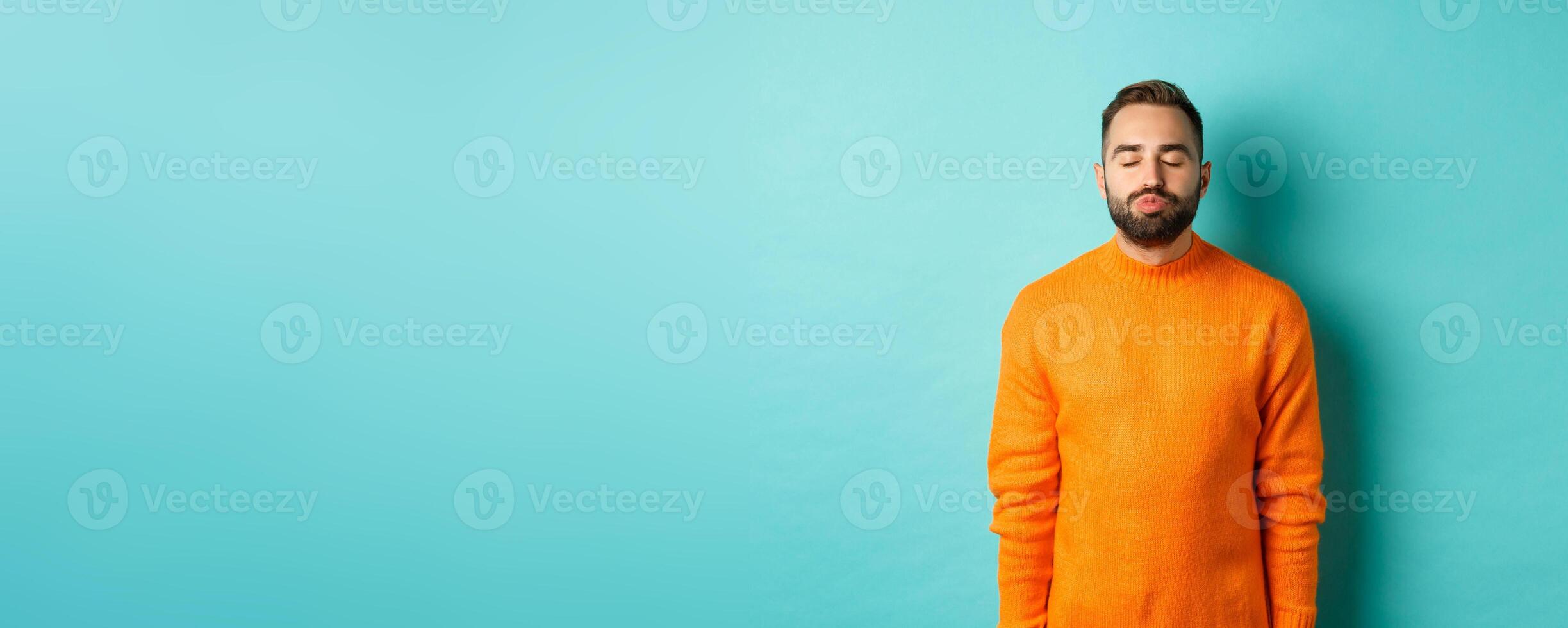 Waist up shot of young man pucker lips and close eyes, waiting for kiss, standing in orange sweater against light blue background photo