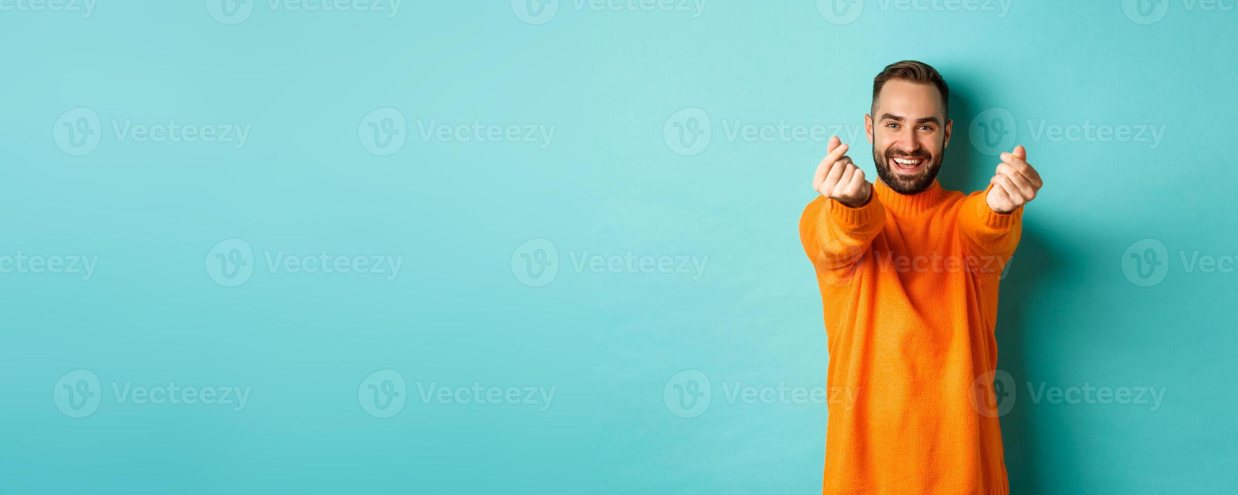 Smiling handsome man showing hearts and looking happy, lucky gesture, standing in orange sweater over light blue background photo