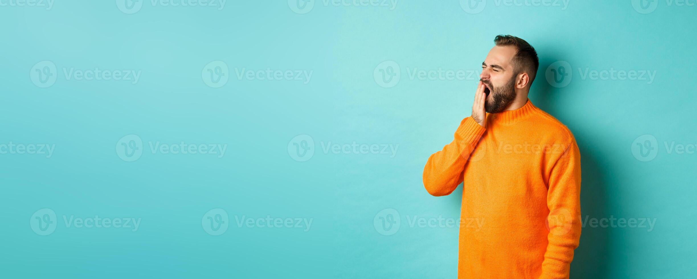 Photo of tired bearded man yawning, cover mouth with hand, standing sleepy against light blue background