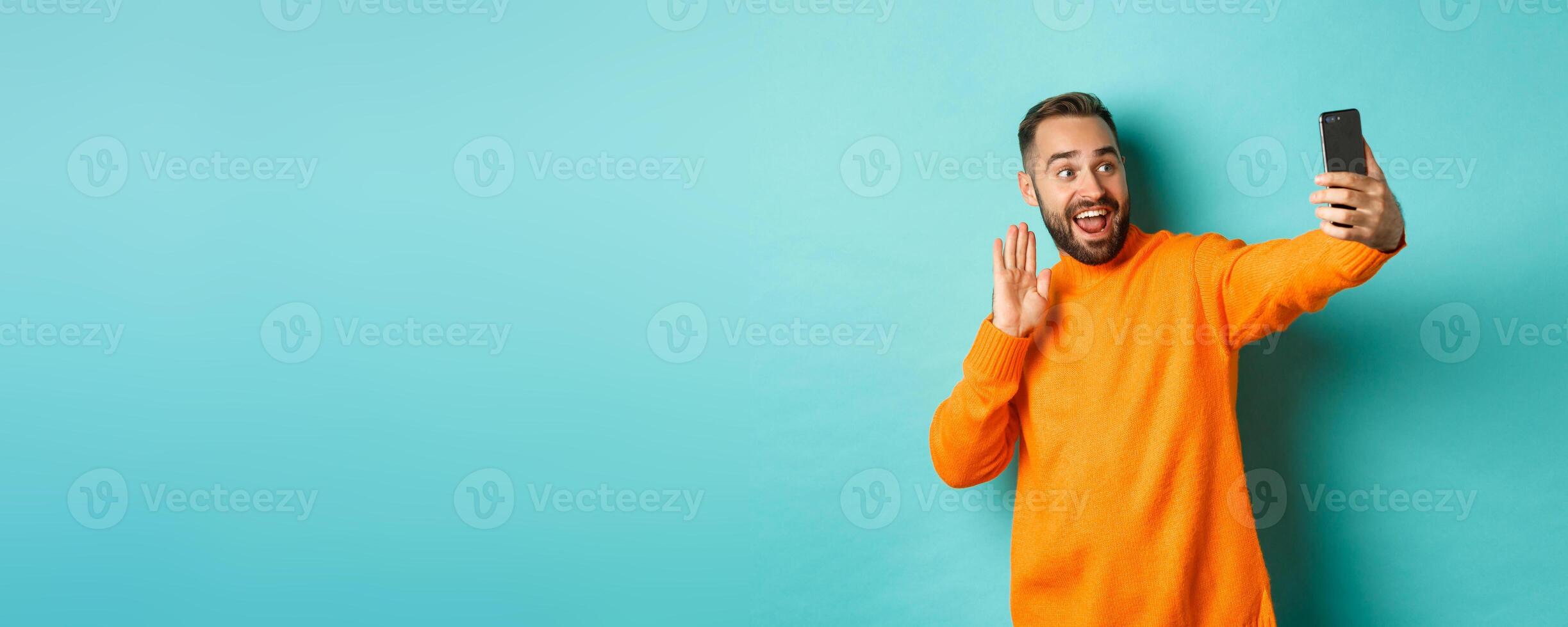 Happy young man calling, talking online with mobile phone, saying hello to smartphone camera and waving hand friendly, standing over light blue background photo