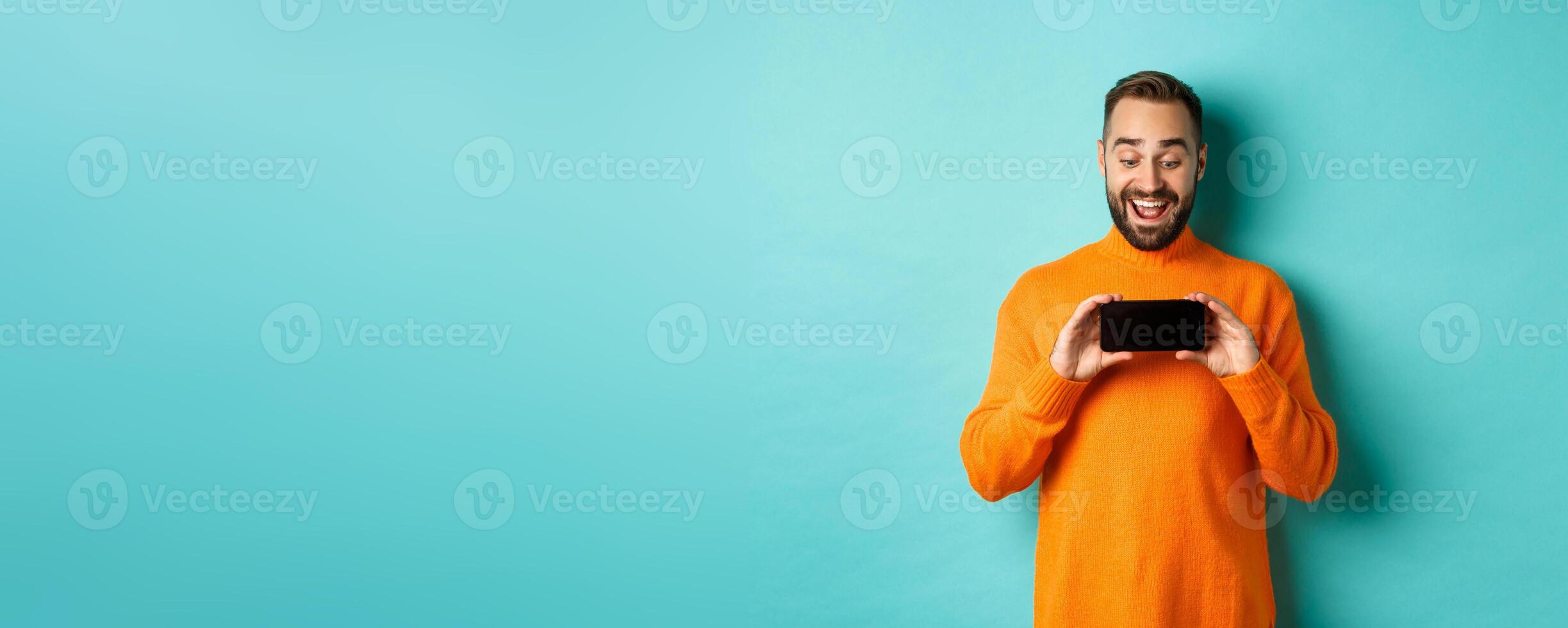 Excited guy demonstrating online store, showing mobile screen and looking amazed, standing over light blue background photo
