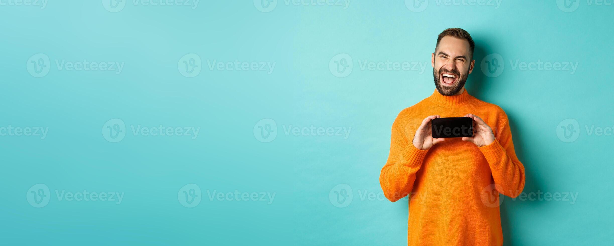 Photo of happy adult man laughing and showing funny thing on mobile phone screen, standing in orange sweater over turquoise background