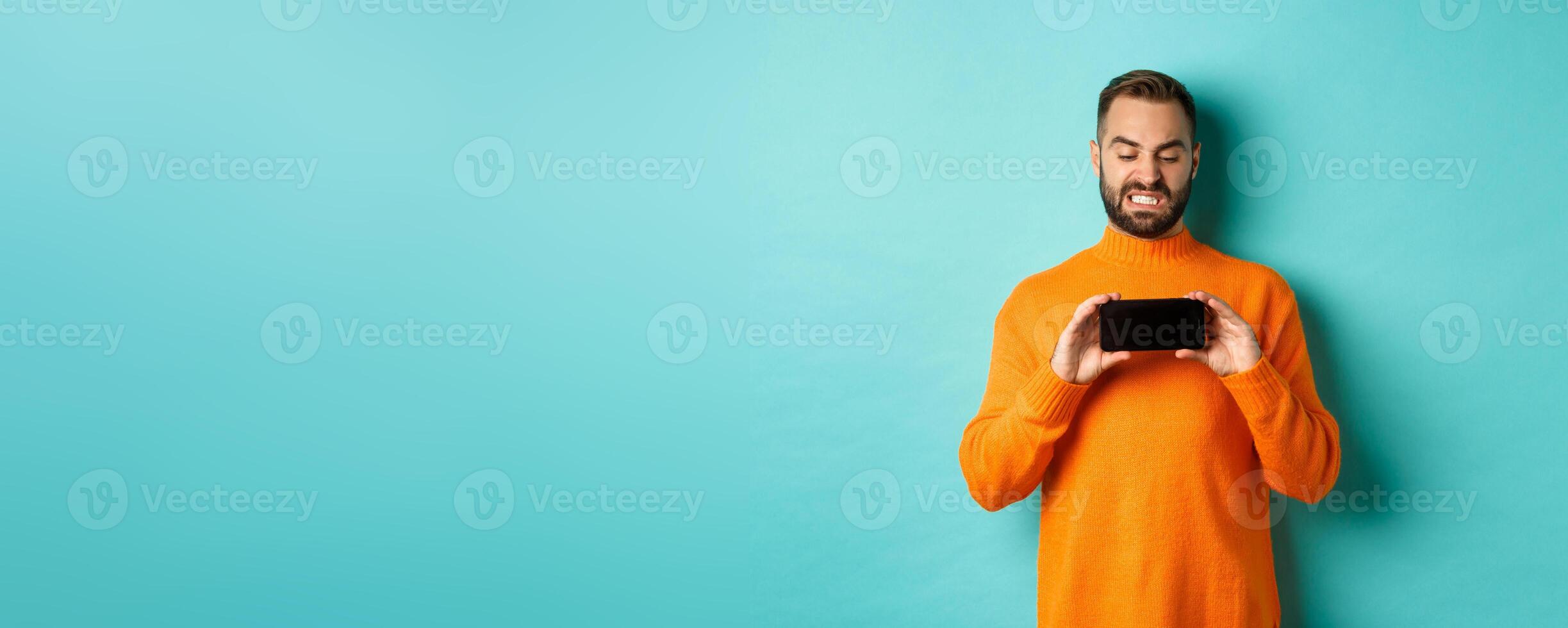 Image of man cringe at something on mobile screen, staring disgusted at display, showing smartphone, standing over light blue background photo