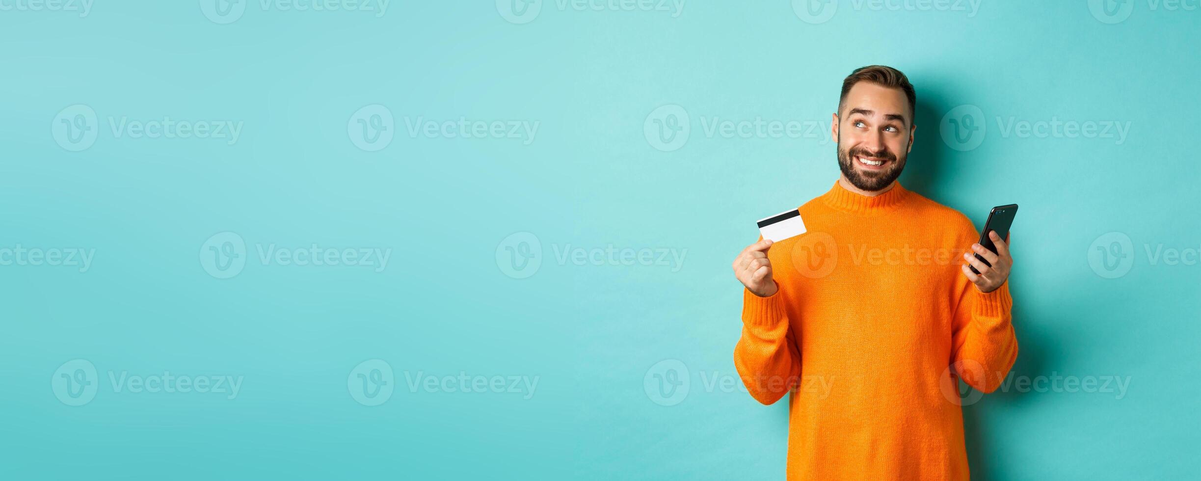 Online shopping. Handsome man thinking, holding smartphone with credit card, paying in internet store, standing over light blue background photo