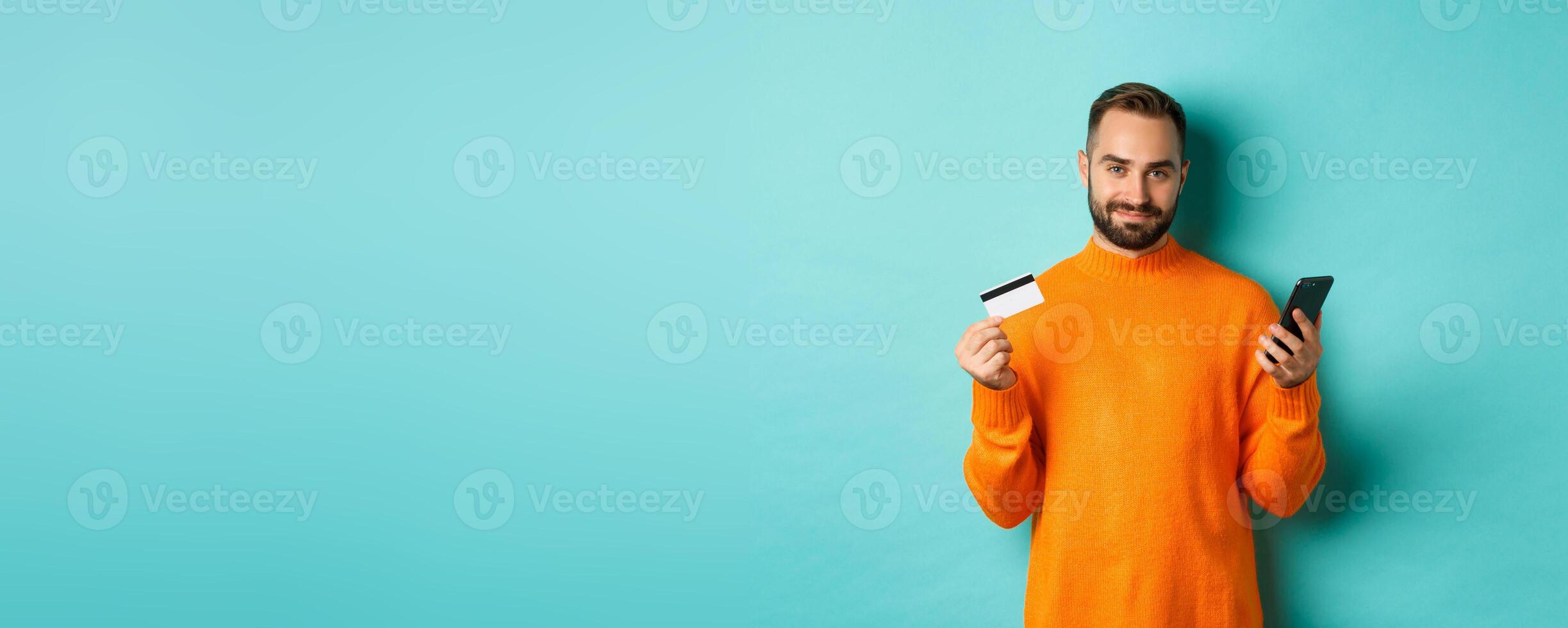 Online shopping. Handsome caucasian man in orange sweater, using credit card and mobile phone, standing over light blue background photo
