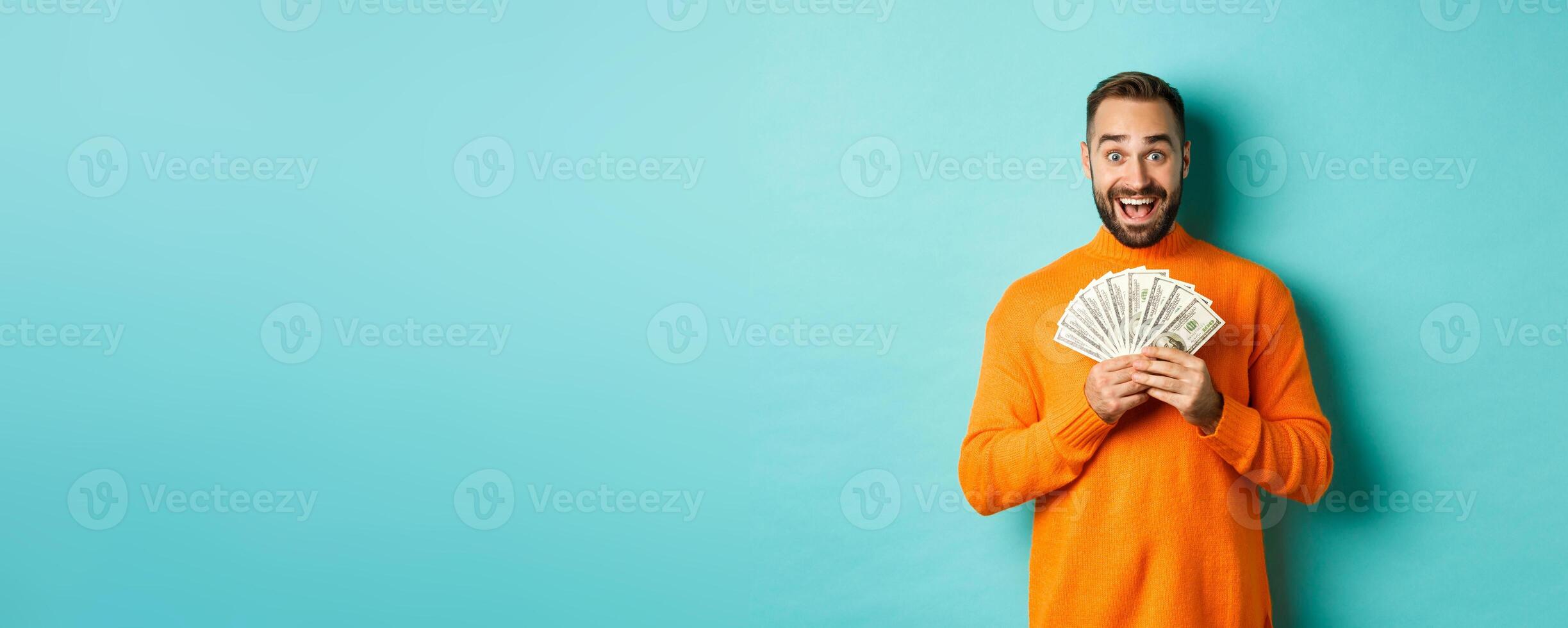 Image of excited bearded guy holding money and rejoicing, winning cash prize, standing against light blue background photo