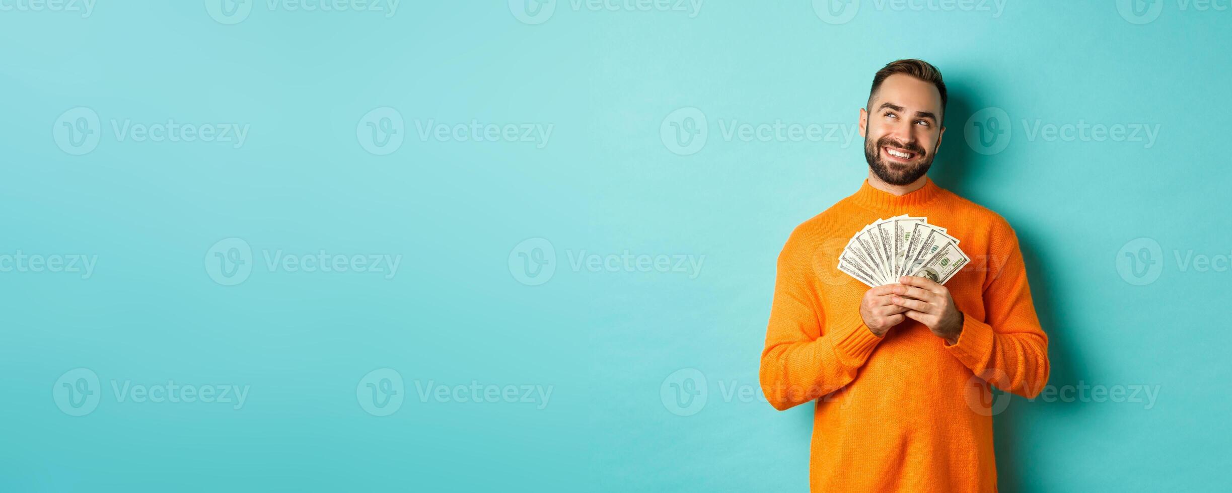 Man thinking about shopping, holding money and smiling satisfied, looking at upper left corner, standing over light blue background photo