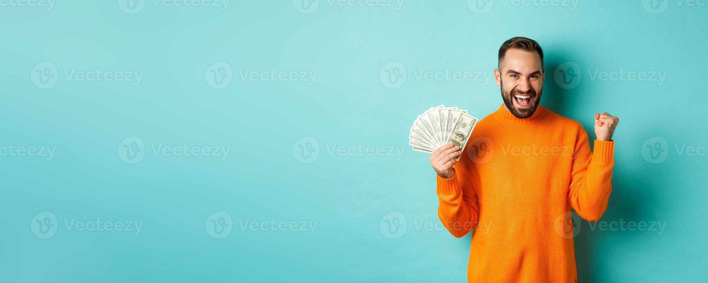 Shopping. Cheerful guy holding money, winning prize in cash and making fist pump, triumphing with satisfied expression, standing in orange sweater against turquoise background photo