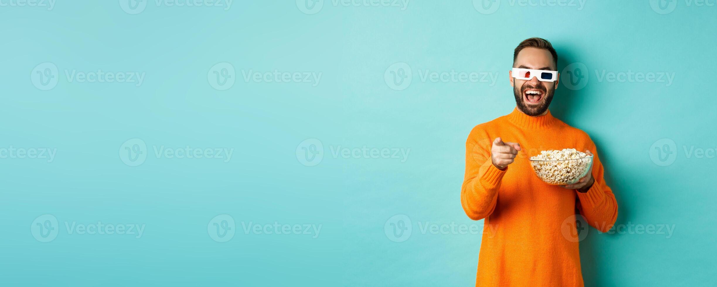 Man watching comedy in 3d glasses, eating popcorn, laughing and pointing at camera tv screen, standing over blue background photo