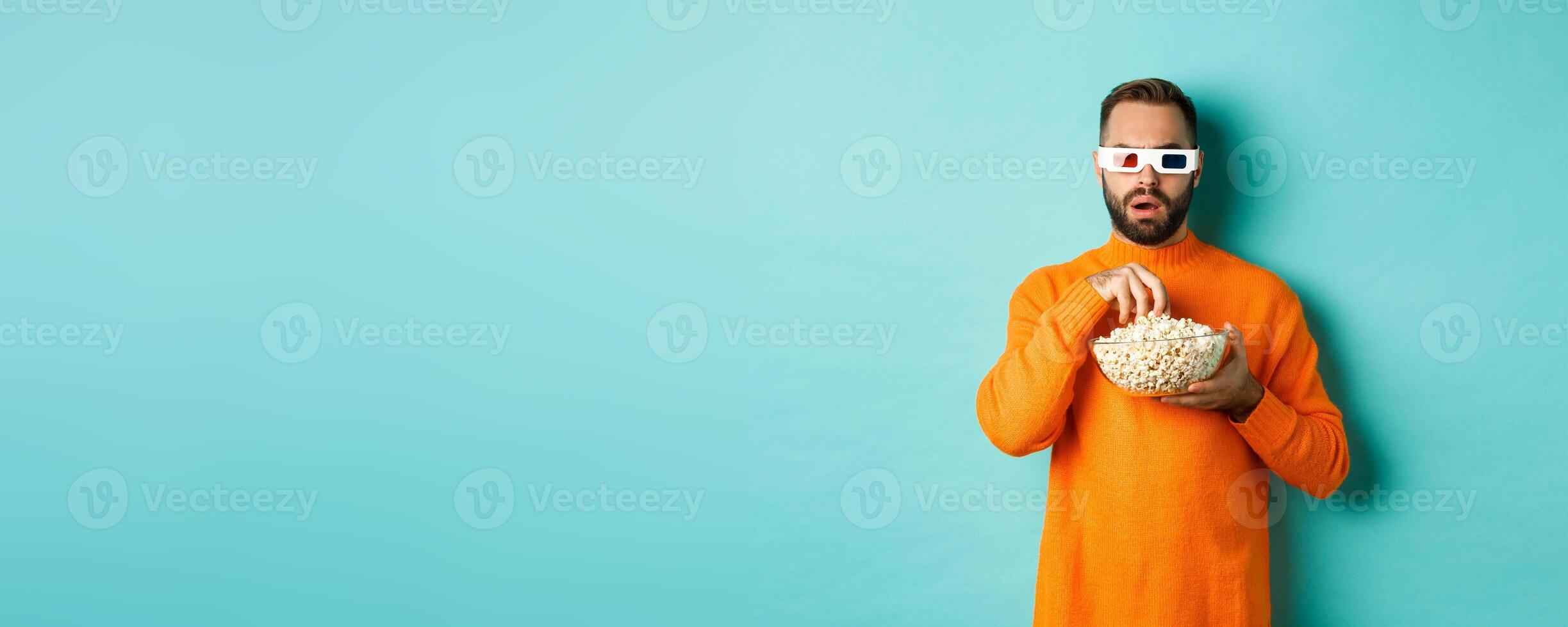 Image of intrigued man eating popcorn and watching movie in 3d, looking at tv screen, standing over blue background photo
